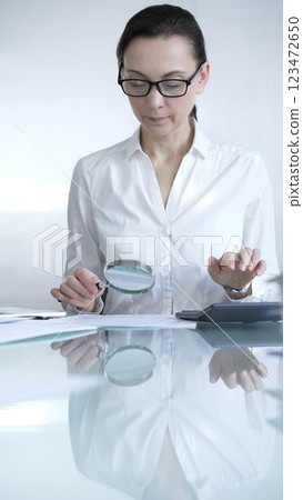 Businesswoman or accountant, wearing white casual shirt and glasses, is using magnifying glass and calculator to analyze financial documents in office setting. Audit and taxes in business 123472650