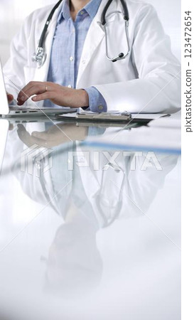 Unrecognizable doctor man in a blue shirt under a medical coat is using technology in medicine. Close-up of hands typing on a laptop on a glass desk in clinic, vertical view. Health care theme concept 123472654