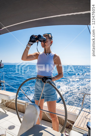 Young female captain on the yacht looking through binoculars during sailing boat control. Travel and active life. Yachting Young female captain on the yacht looking through binoculars during sailing boat control. Travel and active life. Yachting 123476054