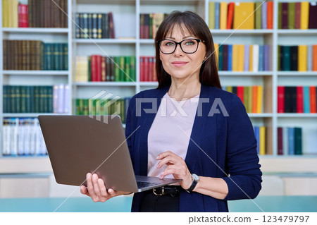 Portrait of middle aged smiling woman teacher professor with laptop inside library 123479797