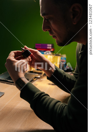 Vertical side view shot of focused technician repairing smartwatch battery, using fine tweezers for precise component extraction and replacement in dark workshop. Vertical side view shot of focused technician repairing smartwatch battery, using fine tweezers for precise component extraction and replacement in dark workshop. 123481067