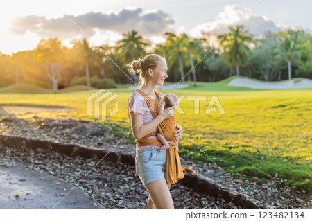 A loving mother walks through a peaceful park, carrying her baby securely in a sling. A heartwarming moment of maternal bonding, babywearing, and the joy of motherhood in nature 123482134