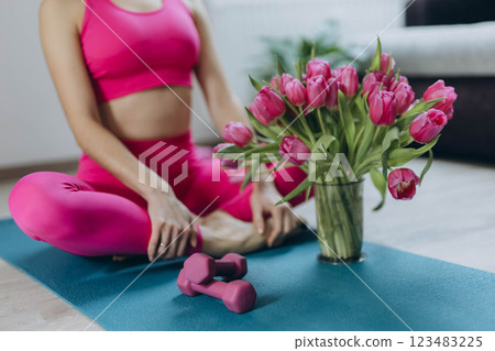 A young woman in a pink sports outfit sits in a lotus pose on a yoga mat, with a pink tulip bouquet 123483225