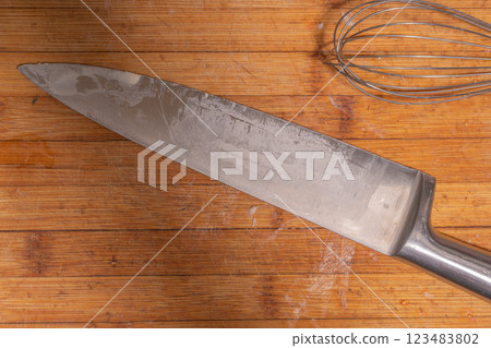 A large kitchen knife and a metal whisk placed on a wooden cutting board. A large kitchen knife and a metal whisk placed on a wooden cutting board. 123483802