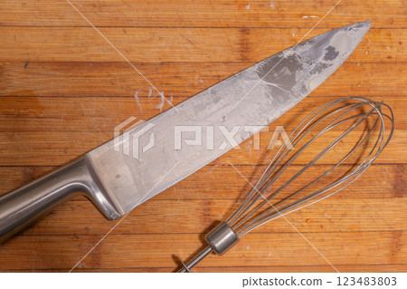 A large kitchen knife and a metal whisk placed on a wooden cutting board. A large kitchen knife and a metal whisk placed on a wooden cutting board. 123483803