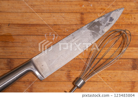 A large kitchen knife and a metal whisk placed on a wooden cutting board. 123483804