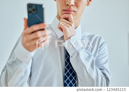 A young man in his twenties holding a smartphone with his hand on his chin 123485215
