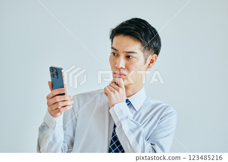A young man in his twenties holding a smartphone with his hand on his chin 123485216