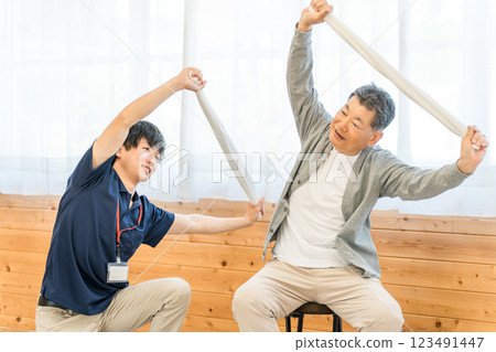 一位老年男性在養老院進行毛巾運動、復健鍛煉，物理治療師、護理員和教練都在場。 123491447