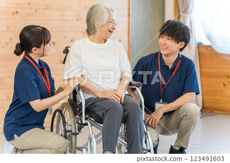 Male and female caregivers and care managers smilingly talk to a senior female patient in a wheelchair (dementia/rehabilitation) Male and female caregivers and care managers smilingly talk to a senior female patient in a wheelchair (dementia/rehabilitation) 123491603