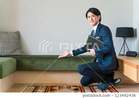 A young male salesman from a house builder wearing a suit shows and proposes sofas in the living room of a model house. 123492436