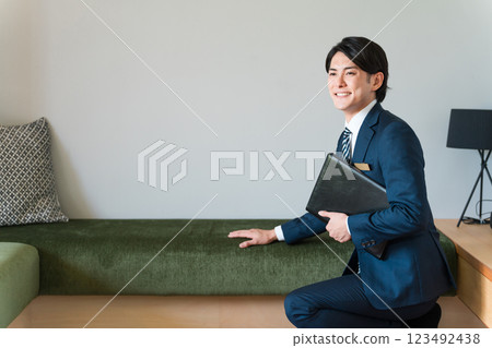 A young male salesman from a house builder wearing a suit shows and proposes sofas in the living room of a model house. A young male salesman from a house builder wearing a suit shows and proposes sofas in the living room of a model house. 123492438