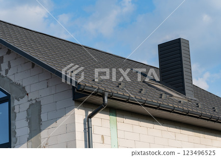 The new house features a contemporary tiled roof with a sleek design, complemented by a prominent chimney. The structure showcases a blend of modern architecture and traditional roofing 123496525