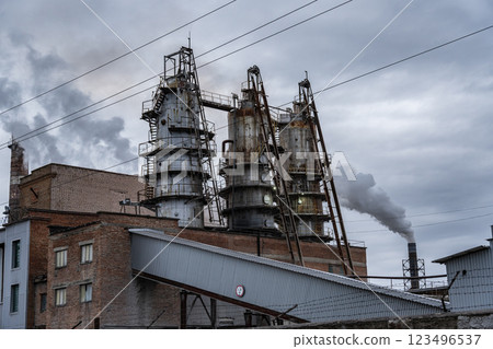 Thick, dark smoke ascends from the high smokestacks of an industrial complex, surrounded by overcast skies and powered machinery, showing signs of activity 123496537