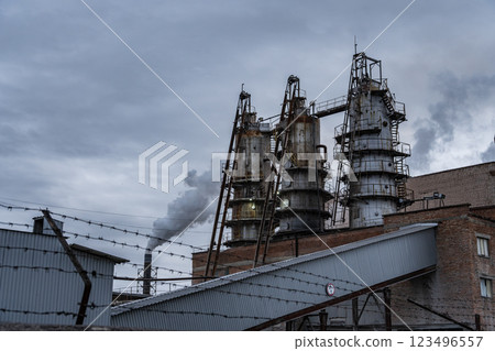 Thick smoke billows from the smokestacks of a manufacturing facility, contrasting against a cloudy sky. The industrial complex is surrounded by barbed wire fencing 123496557