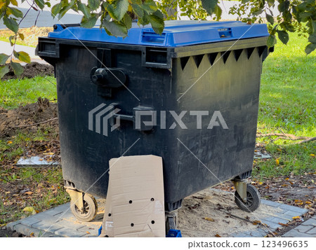 A large, black trash can with a blue lid and wheels is positioned on the edge of a paved street. Grass and fallen leaves are visible in the background 123496635
