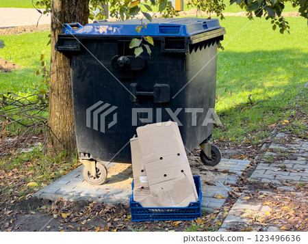 The black trash can sits near a tree on a street, with a cardboard box placed beside it, suggesting recent rubbish collection in a bright, outdoor environment The black trash can sits near a tree on a street, with a cardboard box placed beside it, suggesting recent rubbish collection in a bright, outdoor environment 123496636