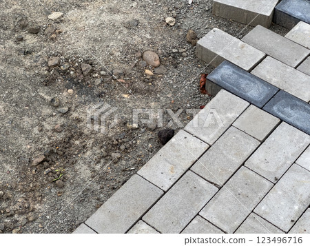 A section of pavement is being constructed, showcasing gray interlocking bricks. Nearby, an unfinished area of gravel and loose stones is visible, indicating progress in development 123496716