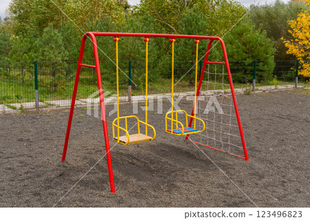 Two brightly colored swings sit in a park with soft ground cover, surrounded by lush greenery, perfect for children to play and enjoy a sunny day outdoors 123496823