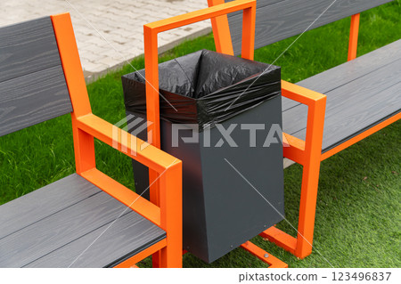 In a vibrant park, a sleek garbage container is mounted between two orange benches, encouraging visitors to dispose of trash responsibly in a clean environment 123496837