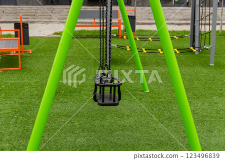 A bright green swing hangs motionless in a playground, inviting children to play while surrounded by lush grass and modern play equipment under a clear sky 123496839