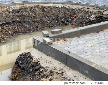 An unfinished brick pavement stretches alongside a sidewalk, featuring a trench filled with water. Loose soil and uninstalled pavers indicate ongoing construction activity 123496869