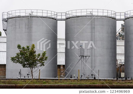 Three massive fuel storage silos rise prominently against a muted background, showcasing industrial architecture with hints of nature nearby, under an overcast sky 123496969