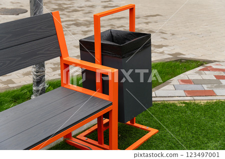 The striking orange bench designed with an attached black trash bin offers convenience for park visitors, enhancing the outdoor experience in a lively setting The striking orange bench designed with an attached black trash bin offers convenience for park visitors, enhancing the outdoor experience in a lively setting 123497001