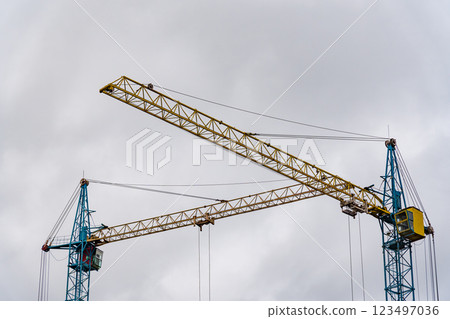Two large construction cranes are actively positioned at a construction site, lifting materials as workers prepare the framework for a new house. The sky above is overcast 123497036