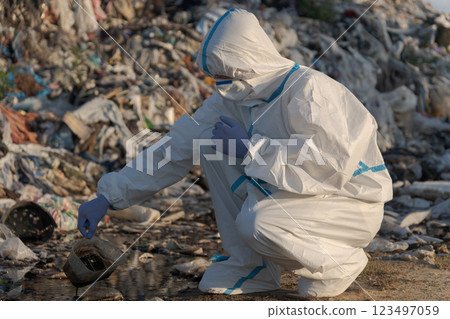 A dedicated environmentalist investigates water pollution at a waste dump site, carefully collecting samples while dressed in protective clothing during late afternoon light A dedicated environmentalist investigates water pollution at a waste dump site, carefully collecting samples while dressed in protective clothing during late afternoon light 123497059