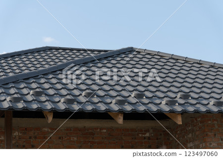 A sleek and modern roofing structure presents grey tiles reflecting the sunlight. This ongoing construction highlights craftsmanship and attention to detail, set against a vibrant blue sky 123497060