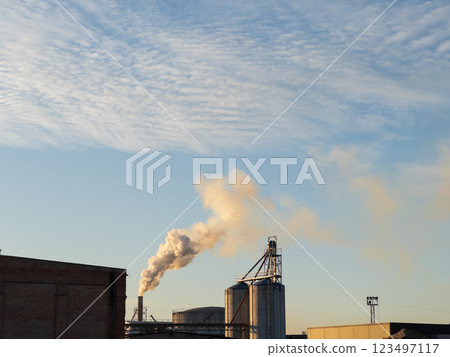 As the sun sets, smoke drifts from large industrial silos, painting a dramatic picture against the expansive blue sky. The quiet evening air hints at a bustling production nearby As the sun sets, smoke drifts from large industrial silos, painting a dramatic picture against the expansive blue sky. The quiet evening air hints at a bustling production nearby 123497117