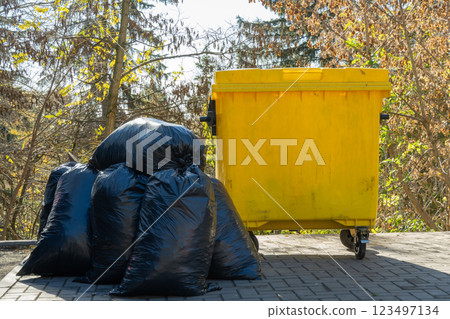 A bright yellow garbage can contrasts against dark plastic bags, set against a backdrop of trees showcasing the colors of fall on a sunny day 123497134