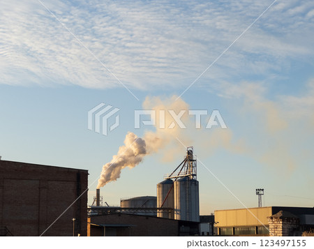 The soft hues of dawn illuminate an industrial area where silos stand tall, releasing gentle clouds of smoke into the crisp morning sky 123497155