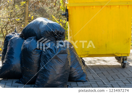 Piles of black garbage bags rest near a vibrant yellow dumpster in a tranquil outdoor area. The setting reveals a serene backdrop of nature, hinting at an upcoming cleanup Piles of black garbage bags rest near a vibrant yellow dumpster in a tranquil outdoor area. The setting reveals a serene backdrop of nature, hinting at an upcoming cleanup 123497274