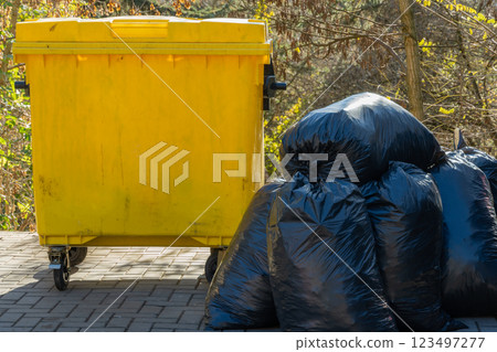 Bright yellow trash bin stands prominently alongside a mound of black garbage bags in a peaceful outdoor location. Sunlight filters through surrounding trees, creating a vivid scene Bright yellow trash bin stands prominently alongside a mound of black garbage bags in a peaceful outdoor location. Sunlight filters through surrounding trees, creating a vivid scene 123497277