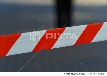 Bright red and white caution tape wraps across the city pavement, serving as a barrier. Its vivid colors stand out under a clear sky, guiding passersby away from a restricted area 123497281