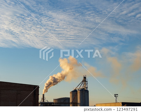 In a sprawling industrial area, smokestacks release plumes of smoke against a backdrop of fluffy clouds. The warm hues of sunset cast a golden light on the structures 123497291