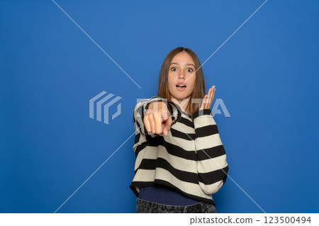 Excited and positive Hispanic teenage girl about 15 years old wearing striped sweater pointing index finger at you and seeing something very funny. Isolated over blue studio background 123500494