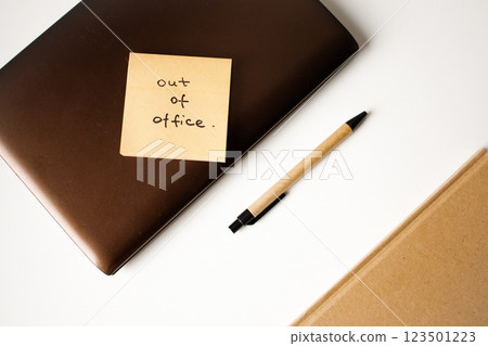 Sticky note with handwritten phrase out of office on a laptop with pen and notebook top view 123501223