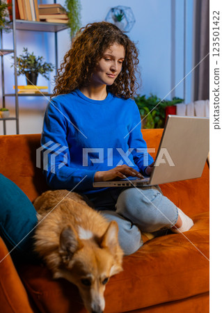 Happy woman opening laptop and working remotely sitting beside corgi dog on sofa at home room 123501422