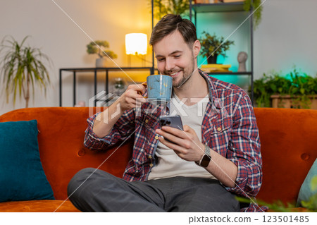 Happy young man uses smartphone having chat on social media while having hot coffee from cup at home 123501485