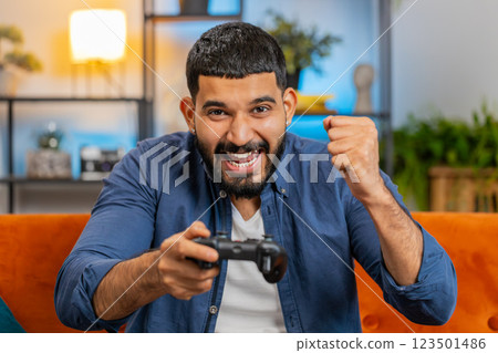 Indian man using joystick controller playing video console winning online fun game sits on home sofa 123501486
