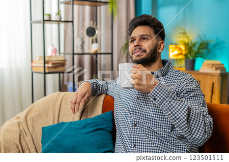 Attractive smiling Indian young man drinking a cup of coffee or herbal tea at home couch in morning Attractive smiling Indian young man drinking a cup of coffee or herbal tea at home couch in morning 123501511