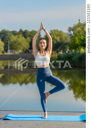 Attractive young woman sportive trainer in activewear standing in yoga Tree Pose near pond in park 123501593