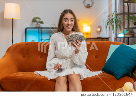 Rich happy young woman counting money cash using smartphone calculator sitting on sofa at home 123501644