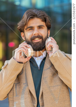 Indian young businessman taking off wireless headphones looking at camera and smiling on city street Indian young businessman taking off wireless headphones looking at camera and smiling on city street 123501977