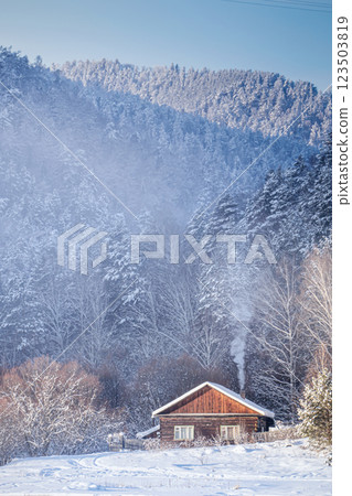 A wooden cottage for a country holiday in a winter forest against the backdrop of high mountains. Smoke from the stove is coming out of the chimney, a warm and cozy atmosphere. A Cozy Cabin Set Within A wooden cottage for a country holiday in a winter forest against the backdrop of high mountains. Smoke from the stove is coming out of the chimney, a warm and cozy atmosphere. A Cozy Cabin Set Within 123503819