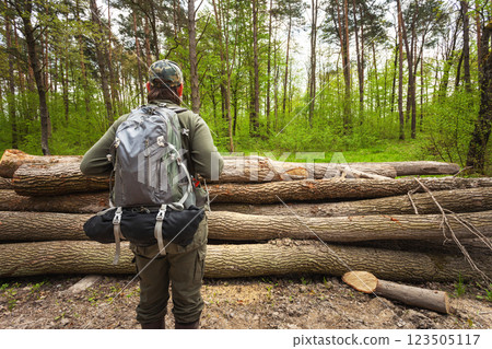 A man with a backpack stands in the forest in front of cut logs. 123505117