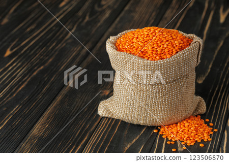 Small burlap sack overflowing with vibrant red lentils, spilling onto a dark, rustic wooden table, creating warm scene 123506870